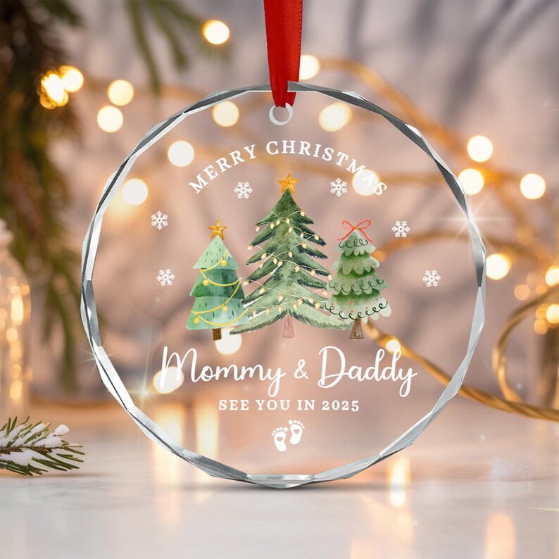 A clear, faceted Christmas ornament with a red ribbon. The ornament features three watercolor Christmas trees, snowflakes, and the text "MERRY CHRISTMAS" and "Mommy & Daddy SEE YOU IN 2025" with baby footprints.