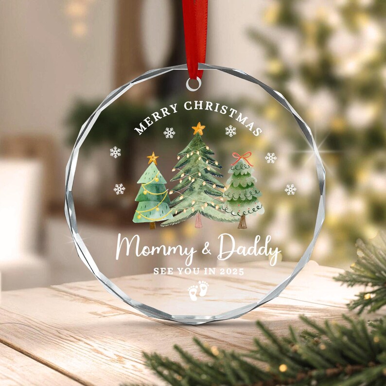 A clear, faceted Christmas ornament with a red ribbon. The ornament features three Christmas trees, snowflakes, and the text "Merry Christmas" and "Mommy & Daddy". The text "See you in 2025" and baby footprints are also visible.