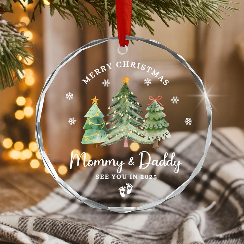 A clear, round Christmas ornament with a red ribbon. The ornament features three watercolor Christmas trees, snowflakes, and the text "MERRY CHRISTMAS", "Mommy & Daddy", and "SEE YOU IN 2025".
