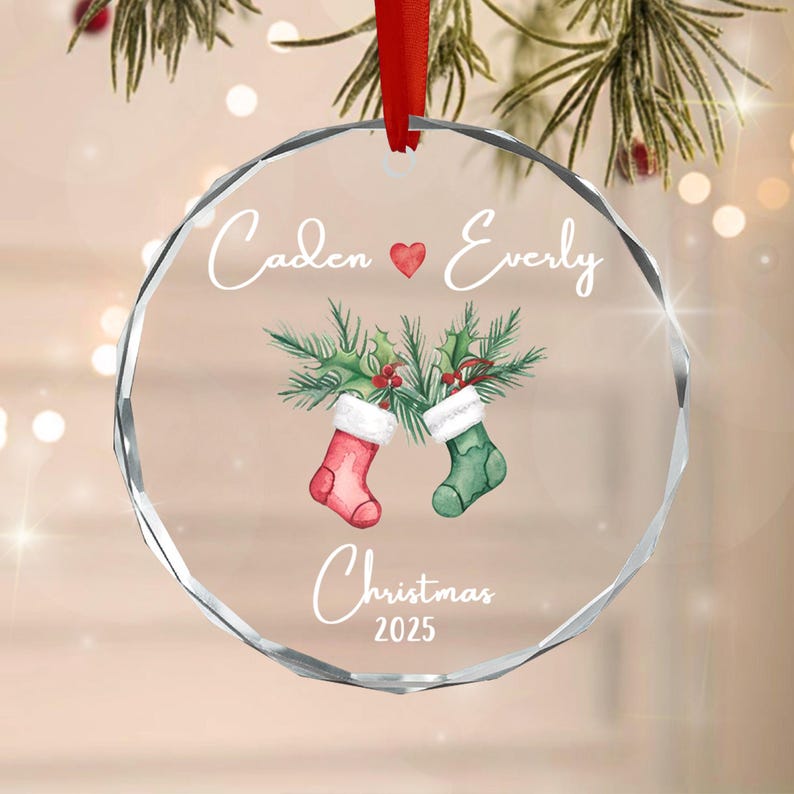 A circular clear glass Christmas ornament with a faceted edge, hanging from a red ribbon. It features the text Caden, a red heart, Everly, and Christmas 2025, with a watercolor illustration of two stockings and holly branches.