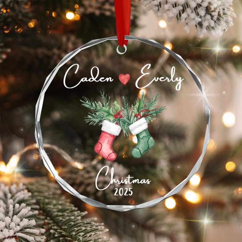 A circular clear acrylic Christmas ornament with faceted edges, featuring the printed names Caden and Everly separated by a red heart, two hanging stockings with holly branches, and the text Christmas 2025.