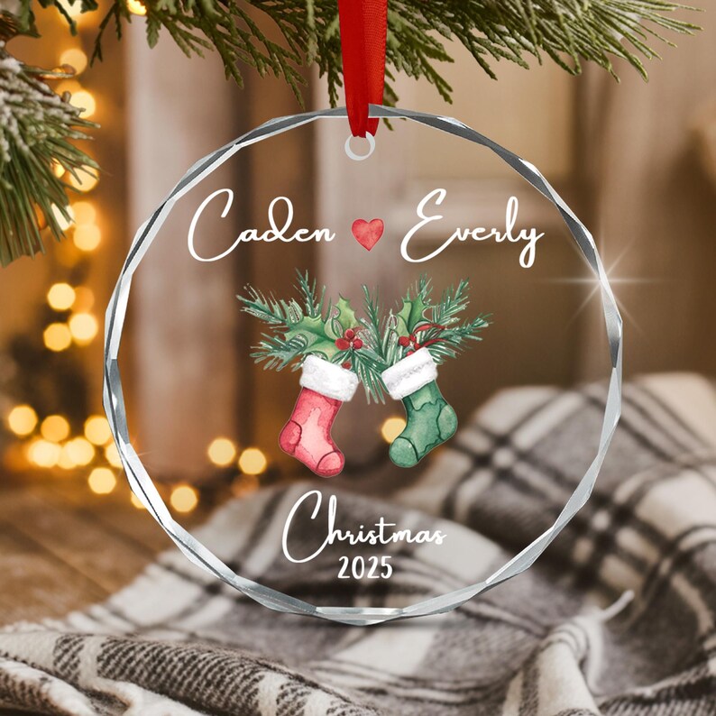 A circular, faceted clear glass ornament hanging from a red ribbon. It features the text Caden and Everly separated by a red heart, a watercolor illustration of two stockings with pine branches, and the text Christmas 2025.