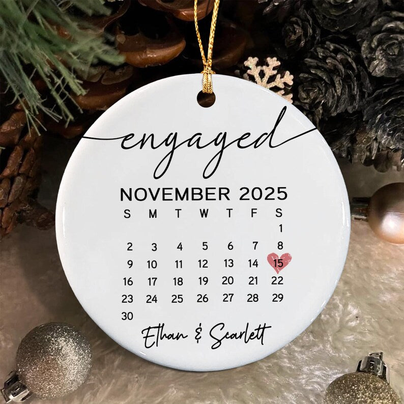 A round white ceramic ornament featuring the word engaged in black script at the top. Below is a November 2025 calendar with a small pink heart over the 15th. The names Ethan and Scarlett are printed in black script at the bottom.