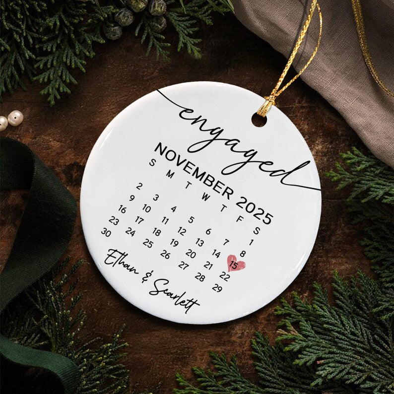 A round white ceramic ornament featuring the word engaged in black script, the calendar for November 2025, and the names Ethan & Scarlett.