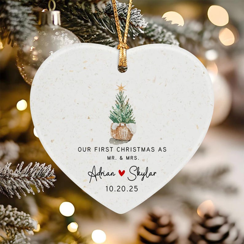 A white heart-shaped ceramic ornament with a gold string. It features a watercolor illustration of a small Christmas tree in a basket, with the text "OUR FIRST CHRISTMAS AS MR. & MRS. Adrian ♥ Skylar 10.20.25" printed in black.