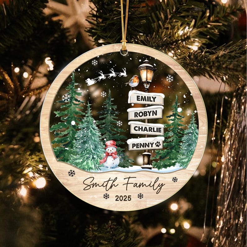 A round wooden Christmas ornament featuring a winter scene with green pine trees, a snowman, a street lamp, and a flying sleigh. The street sign displays the names Emily, Robyn, Charlie, and Penny. Text reads Smith Family 2025.