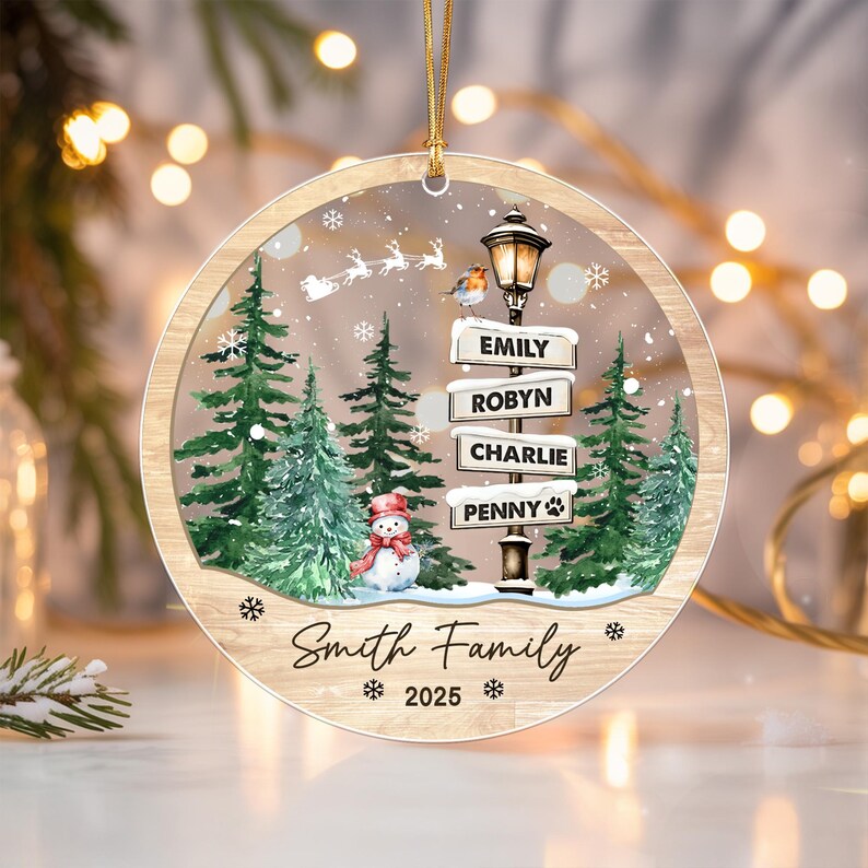 A round, flat Christmas ornament with a wood-grain border. The center features a snowy forest scene with a snowman, a street lamp with signs reading EMILY, ROBYN, CHARLIE, and PENNY, a robin, and Santa's sleigh.