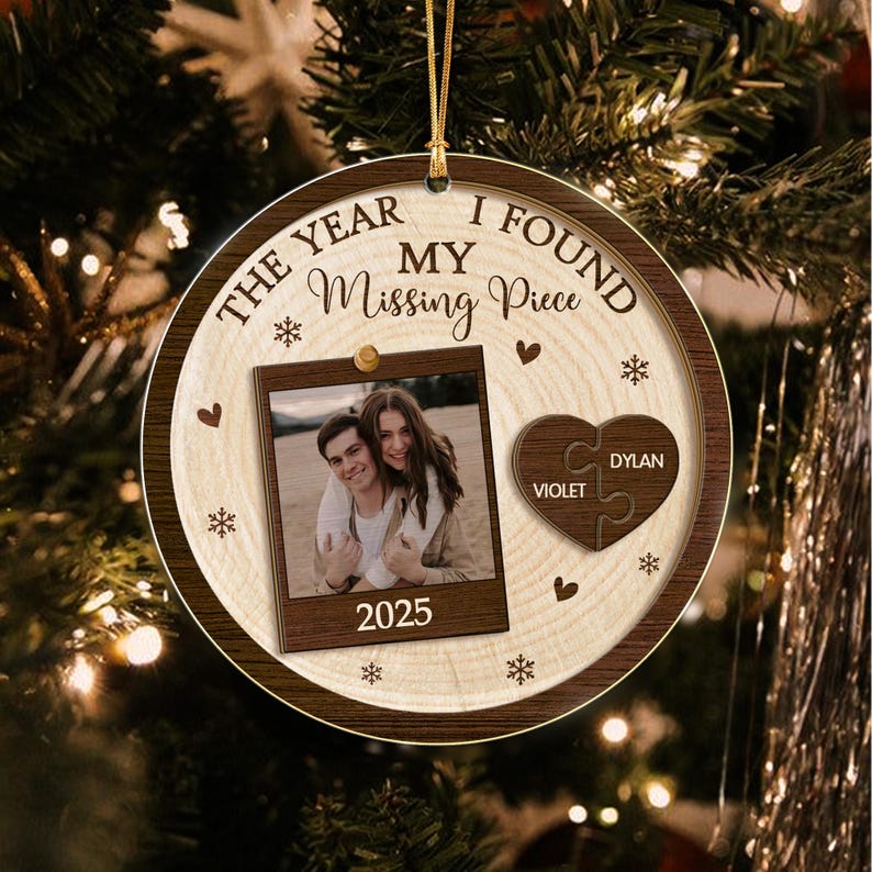 A round wooden Christmas ornament featuring the text THE YEAR I FOUND MY Missing Piece, a photo of a couple, the year 2025, and a heart-shaped puzzle piece with the names VIOLET and DYLAN engraved on the surface.