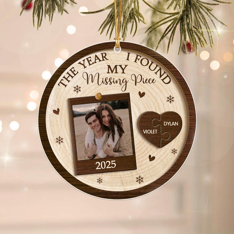 A round wooden Christmas ornament featuring the text THE YEAR I FOUND MY Missing Piece. It includes a central photo frame with a couple, the year 2025, and a heart-shaped puzzle piece ornament with the names VIOLET and DYLAN.