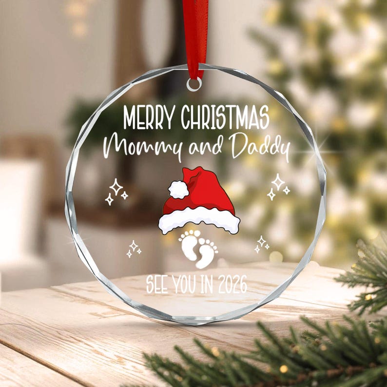 A circular clear glass Christmas ornament with faceted edges, hanging from a red ribbon. It features white text reading Merry Christmas Mommy and Daddy, a red Santa hat, white baby footprints, and the text SEE YOU IN 2026.