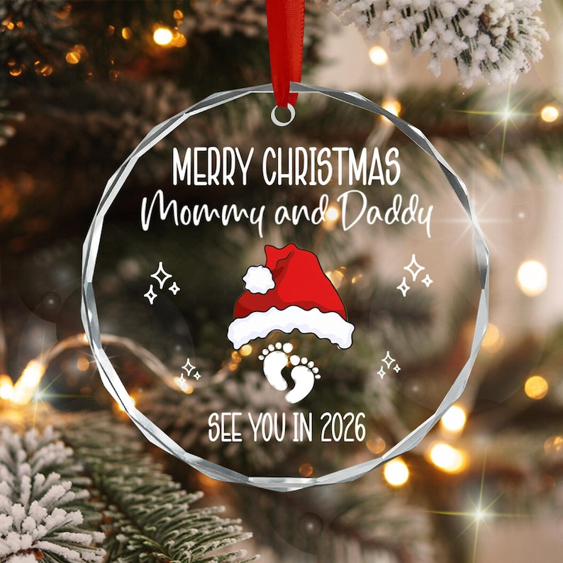 A circular clear glass Christmas ornament with a faceted edge, featuring the white text Merry Christmas Mommy and Daddy, a red and white Santa hat, a pair of baby footprints, and the white text See You In 2026.