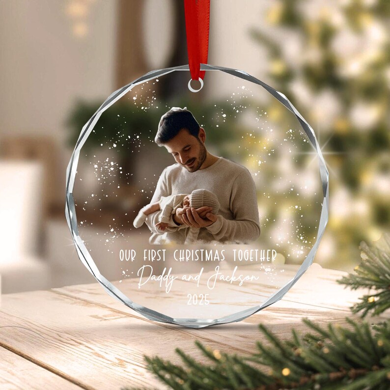 A round, faceted glass Christmas ornament featuring a printed photograph of a person holding a baby. The text reads: OUR FIRST CHRISTMAS TOGETHER, Daddy and Jackson, 2025. A red ribbon is attached to the top for hanging.