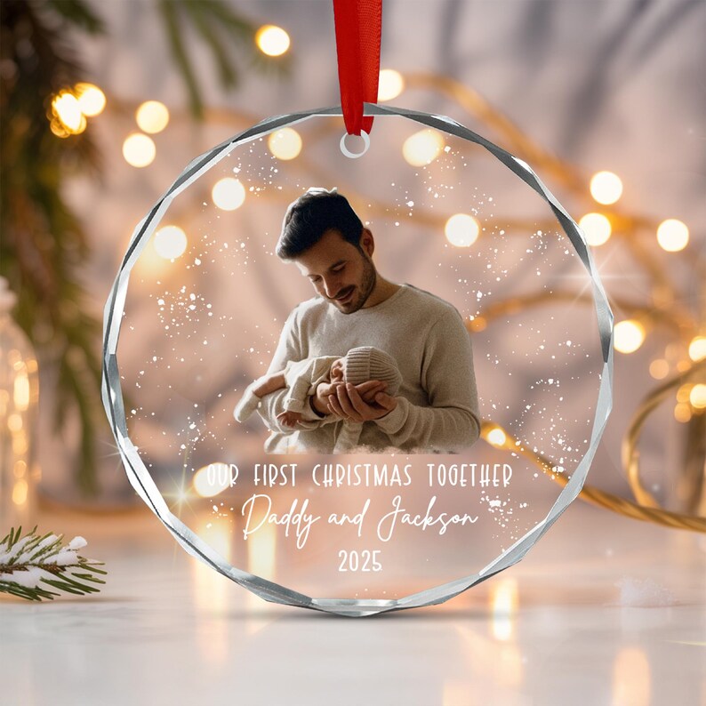 A circular clear glass ornament with faceted edges, featuring a printed photograph of a person holding a baby. The text reads: OUR FIRST CHRISTMAS TOGETHER, Daddy and Jackson, 2025. A red ribbon is attached to the top loop.