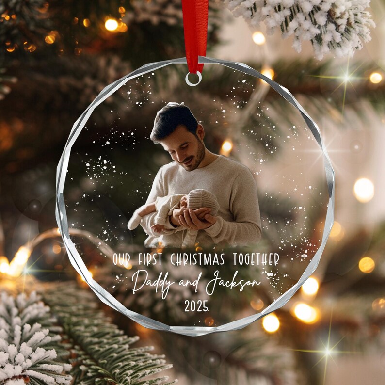 A round, faceted glass Christmas ornament featuring a printed photograph of a person holding a baby. The text reads: OUR FIRST CHRISTMAS TOGETHER, Daddy and Jackson, 2025. A red ribbon is attached to the top for hanging.