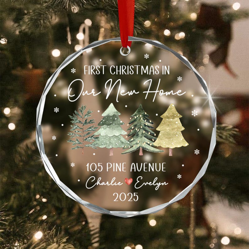 A round, clear glass Christmas ornament with a faceted edge, featuring a red ribbon hanger. The text reads: FIRST CHRISTMAS IN Our New Home, 105 PINE AVENUE, Charlie, a red heart, Evelyn, and 2025.