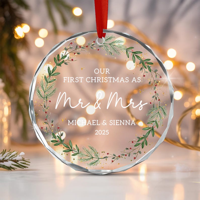 A circular clear acrylic Christmas ornament with faceted edges, featuring a printed green pine wreath with red berries. The text reads: OUR FIRST CHRISTMAS AS. Mr & Mrs MICHAEL & SIENNA 2025. A red ribbon is attached to the top.