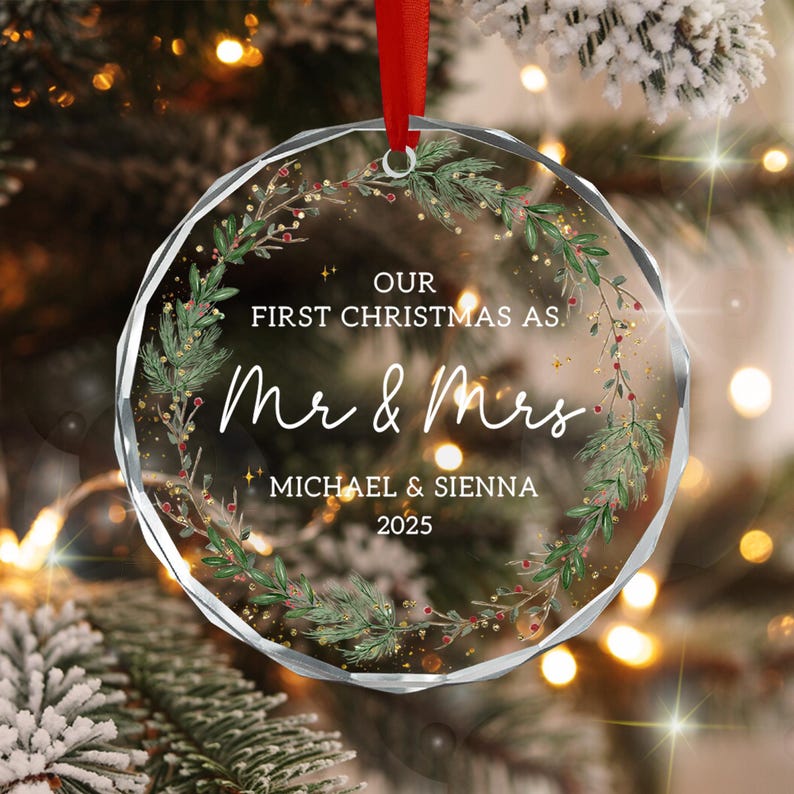 A circular, faceted glass Christmas ornament featuring a printed wreath of green pine branches and red berries. The text reads: OUR FIRST CHRISTMAS AS. Mr & Mrs MICHAEL & SIENNA 2025.