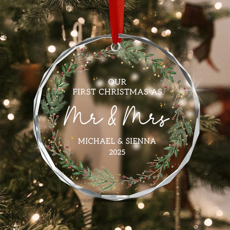 A circular clear glass Christmas ornament with faceted edges, featuring a printed wreath of green pine branches and red berries. The text reads: OUR FIRST CHRISTMAS AS. Mr & Mrs MICHAEL & SIENNA 2025. It hangs from a red ribbon.