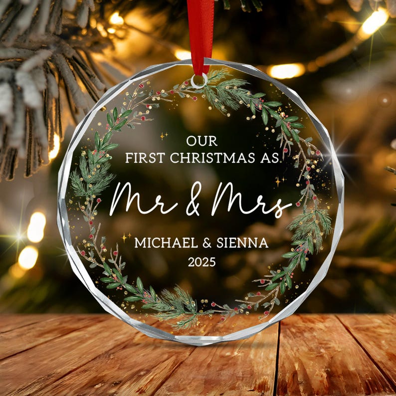A circular, faceted glass Christmas ornament featuring a printed wreath of green pine branches and red berries. The text reads: OUR FIRST CHRISTMAS AS, Mr & Mrs, MICHAEL & SIENNA, 2025.