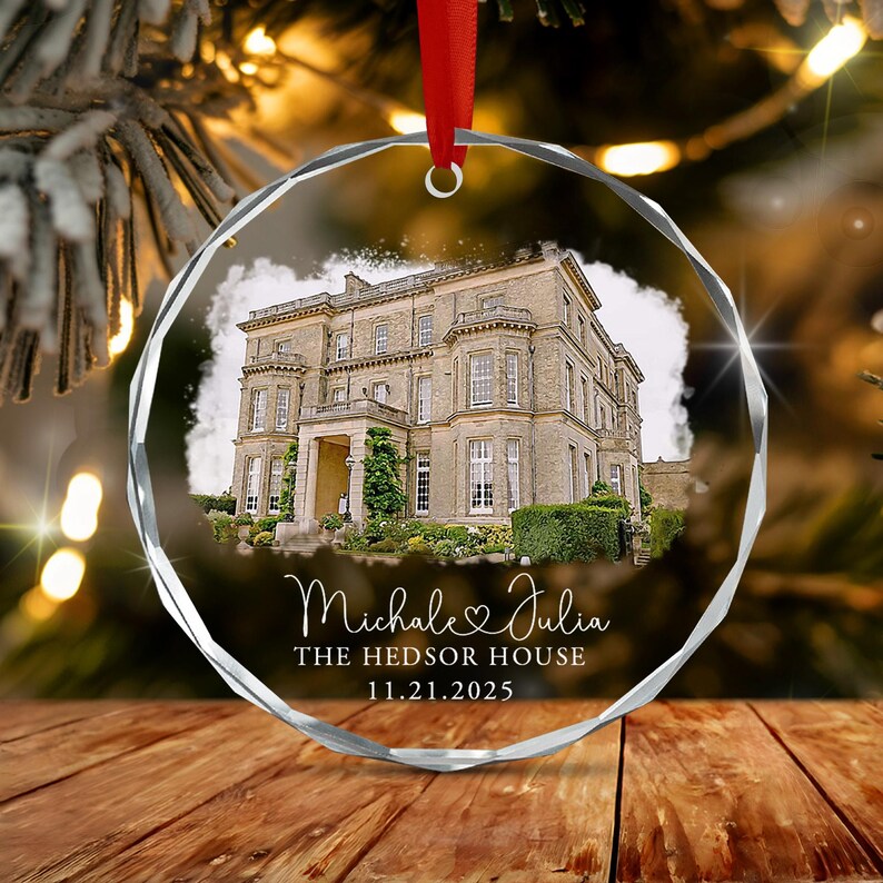 A circular, faceted glass ornament featuring a printed image of a large stone mansion. Below the house, the text reads "Michale & Julia", "THE HEDSOR HOUSE", and "11.21.2025". A red ribbon is attached to the top for hanging.