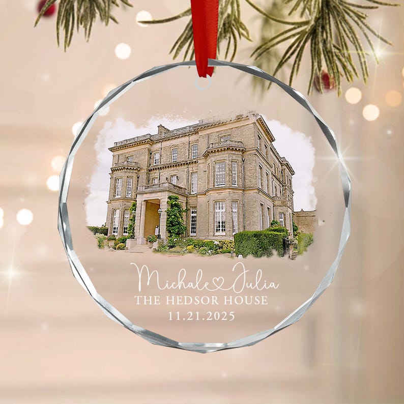 A circular glass ornament featuring a printed image of a large stone mansion. Below the house, the text reads "Michale & Julia", "THE HEDSOR HOUSE", and "11.21.2025". The ornament has a faceted edge and hangs from a red ribbon.