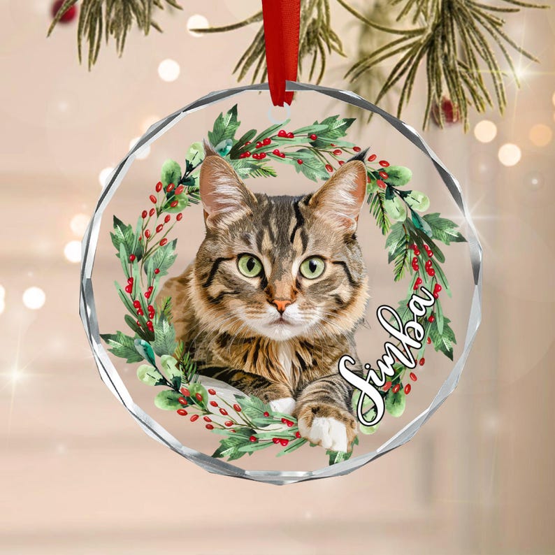 A round, clear acrylic ornament featuring a brown tabby cat centered within a festive wreath of green leaves and red berries. The name Simba is written in elegant black script on the right side.