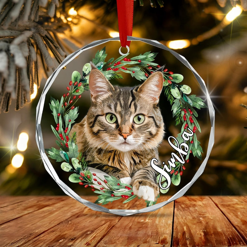 A round, faceted glass ornament featuring a brown tabby cat centered within a wreath of green leaves and red berries. The name Simba is printed in white script on the right side. A red ribbon is attached to the top for hanging.