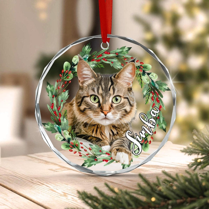 A round, clear acrylic ornament featuring a brown tabby cat centered within a green pine wreath with red berries. The name Simba is printed in white script on the right side. A red ribbon is attached to the top for hanging.