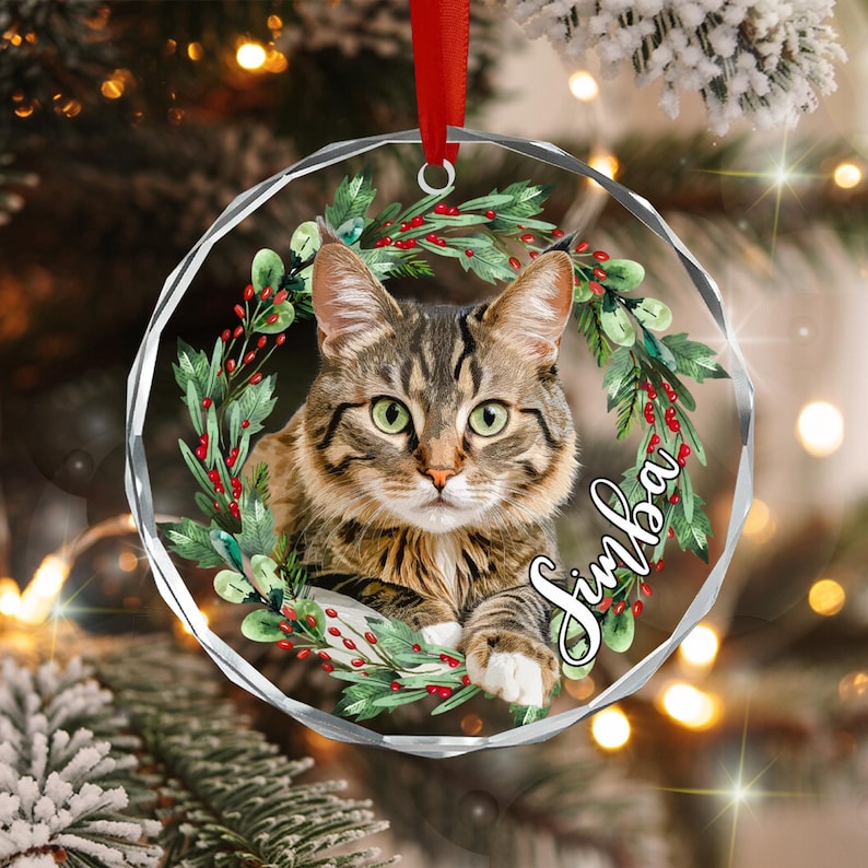 A circular, faceted glass ornament featuring a brown tabby cat centered within a wreath of green pine needles and red berries. The name Simba is printed in white script on the right side. A red ribbon is attached to the top loop.