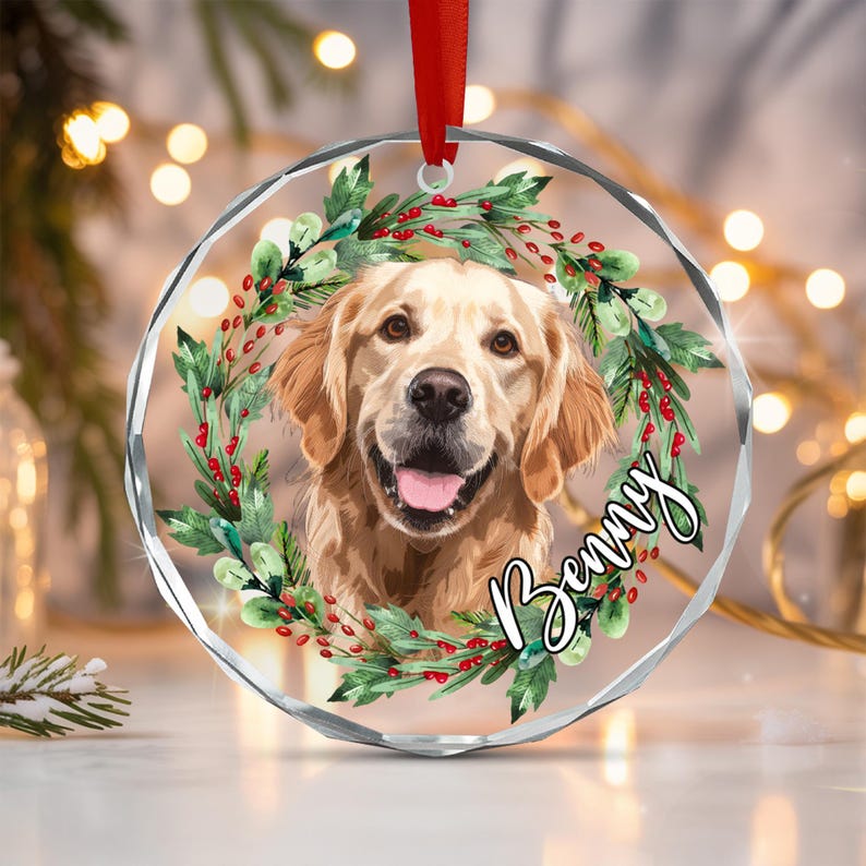 A circular glass ornament featuring a golden retriever portrait surrounded by a wreath of green leaves and red berries. The name Benny is written in white script across the bottom right. A red ribbon is attached to the top.