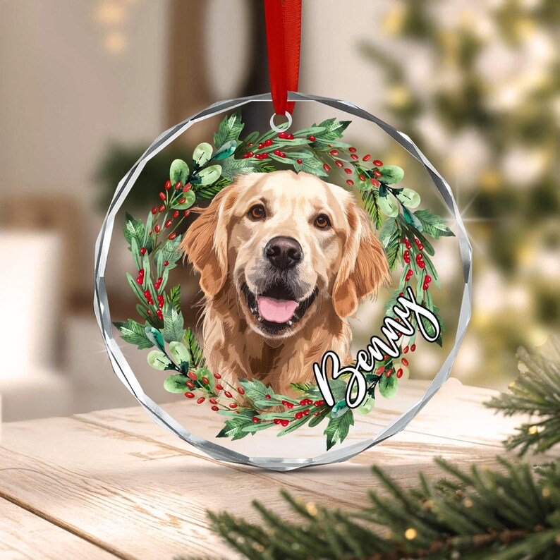 A circular clear acrylic ornament featuring a golden retriever portrait centered within a wreath of green leaves and red berries. The name Benny is written in black script on the right side. A red ribbon is attached to the top.
