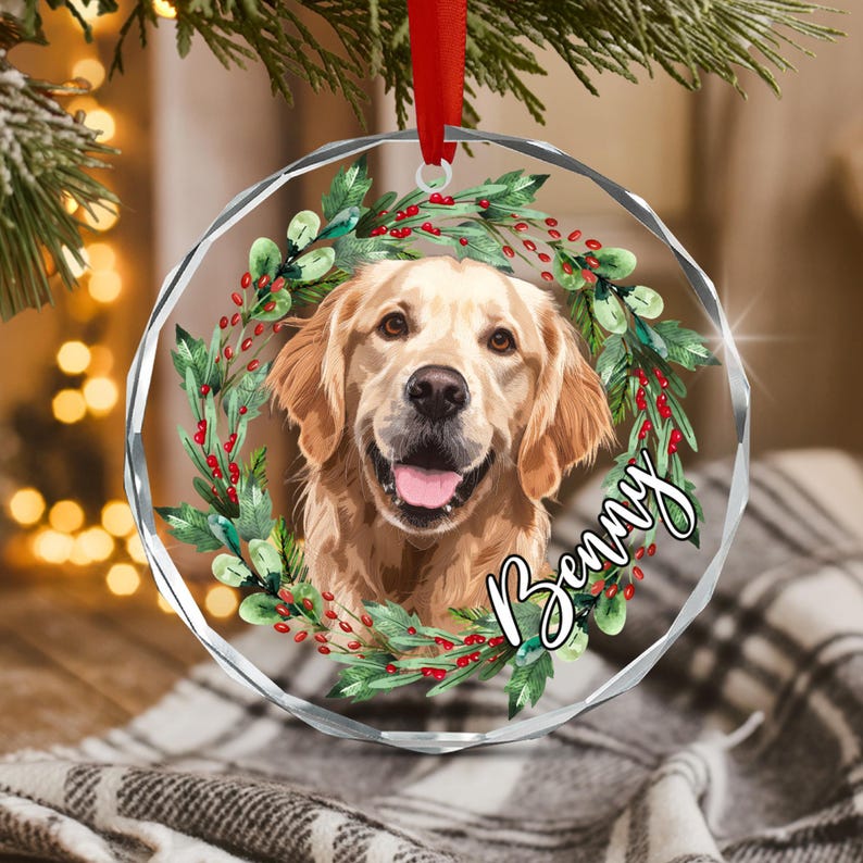 A circular clear acrylic ornament featuring a golden retriever portrait surrounded by a wreath of green leaves and red berries. The name Benny is written in white cursive script on the right side. A red ribbon hangs from the top.