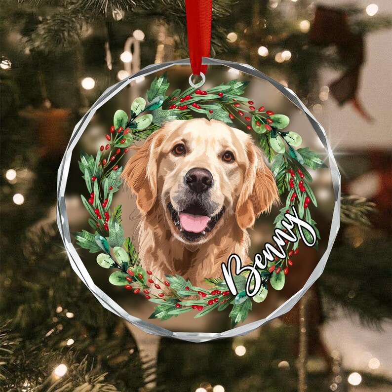 A round, clear acrylic Christmas ornament featuring a golden retriever portrait framed by a green wreath with red berries. The name Benny is printed in white script on the right side. A red ribbon is attached to the top.