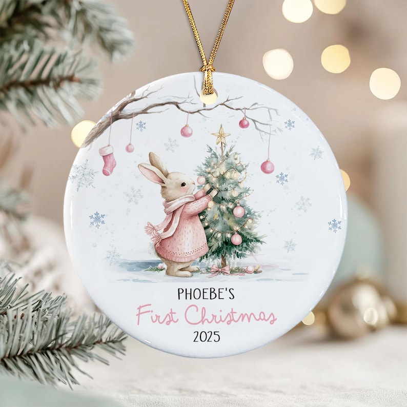 A round white ceramic ornament featuring a watercolor illustration of a rabbit in a pink sweater decorating a Christmas tree. The text reads PHOEBE'S First Christmas 2025.