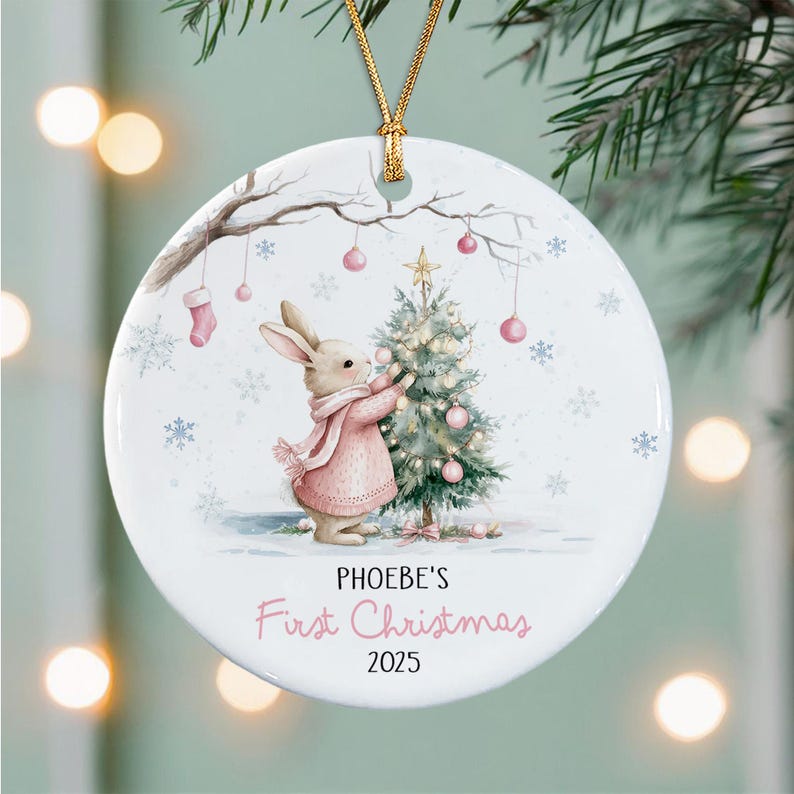 A round white ceramic Christmas ornament featuring a watercolor illustration of a rabbit in a pink sweater decorating a small tree. The text reads PHOEBE'S First Christmas 2025.
