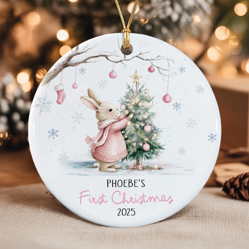 A round white ceramic ornament featuring a watercolor illustration of a rabbit in a pink sweater decorating a Christmas tree. The text reads PHOEBE'S First Christmas 2025.