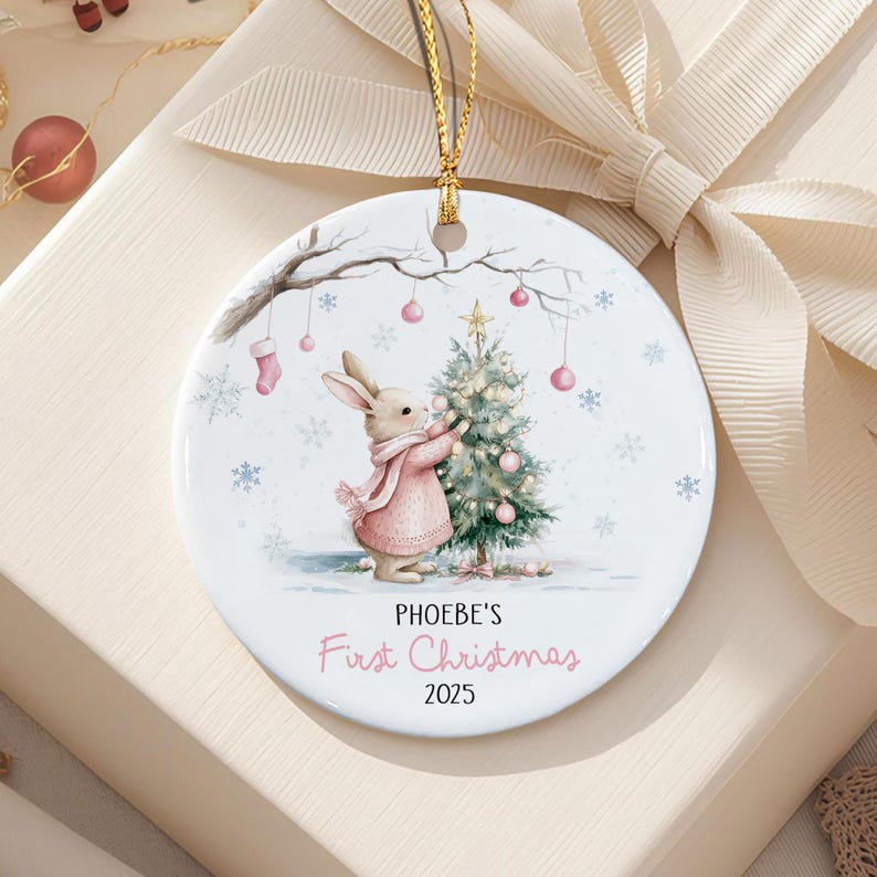 A round white ceramic ornament featuring a watercolor illustration of a rabbit in a pink scarf decorating a Christmas tree. The text reads PHOEBE'S First Christmas 2025.