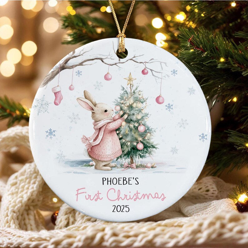 A round white ceramic Christmas ornament featuring a watercolor illustration of a rabbit in a pink sweater decorating a small pine tree. The text reads PHOEBE'S First Christmas 2025 in black and pink script.