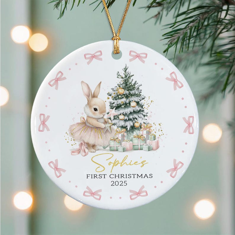 A round white ceramic ornament hanging from a gold cord. The design features a rabbit in a tutu standing next to a decorated Christmas tree with gifts.