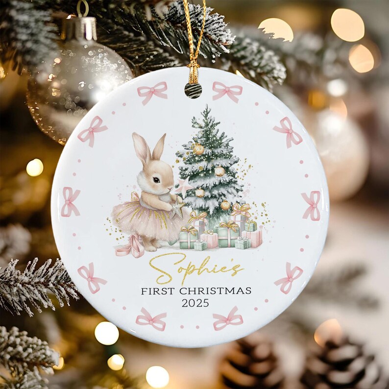 A round white ceramic ornament featuring a bunny in a tutu standing next to a decorated Christmas tree with gifts. The text reads Sophie's FIRST CHRISTMAS 2025. The border is decorated with small pink bows and dots.