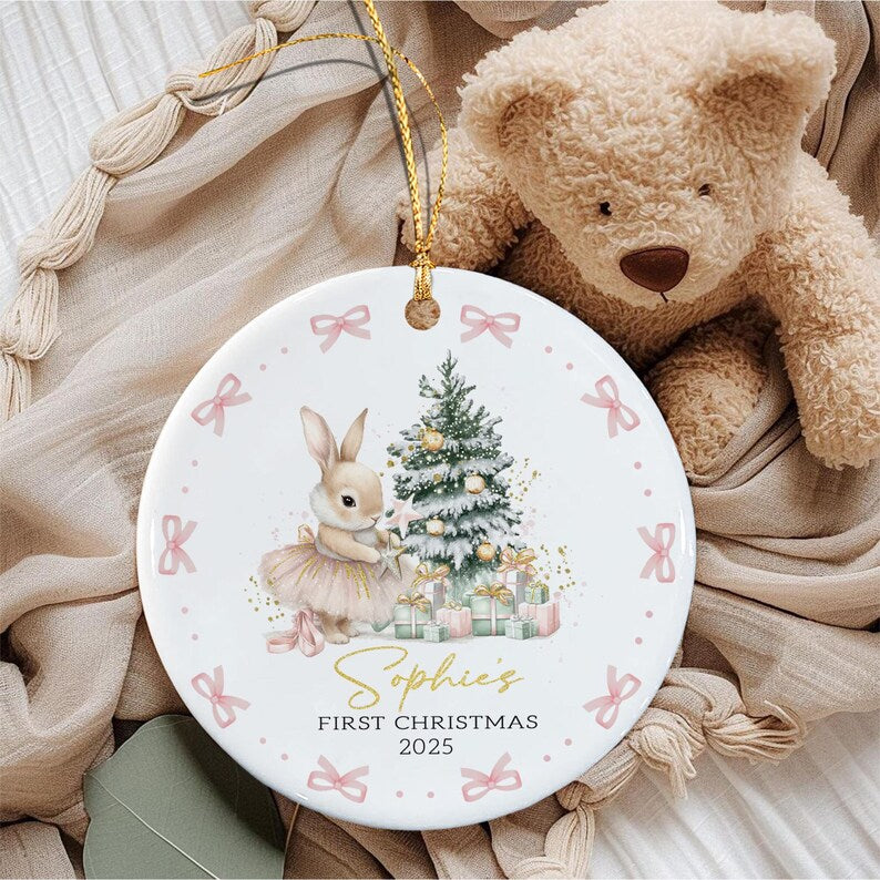 A round white ceramic ornament featuring a bunny in a tutu standing next to a decorated Christmas tree with gifts. The text reads Sophie's FIRST CHRISTMAS 2025.