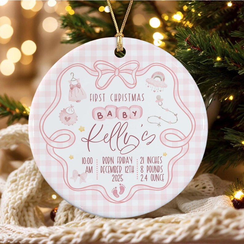 A round white ceramic ornament with a pink gingham pattern, featuring a pink bow, baby clothes, and a pacifier graphic.