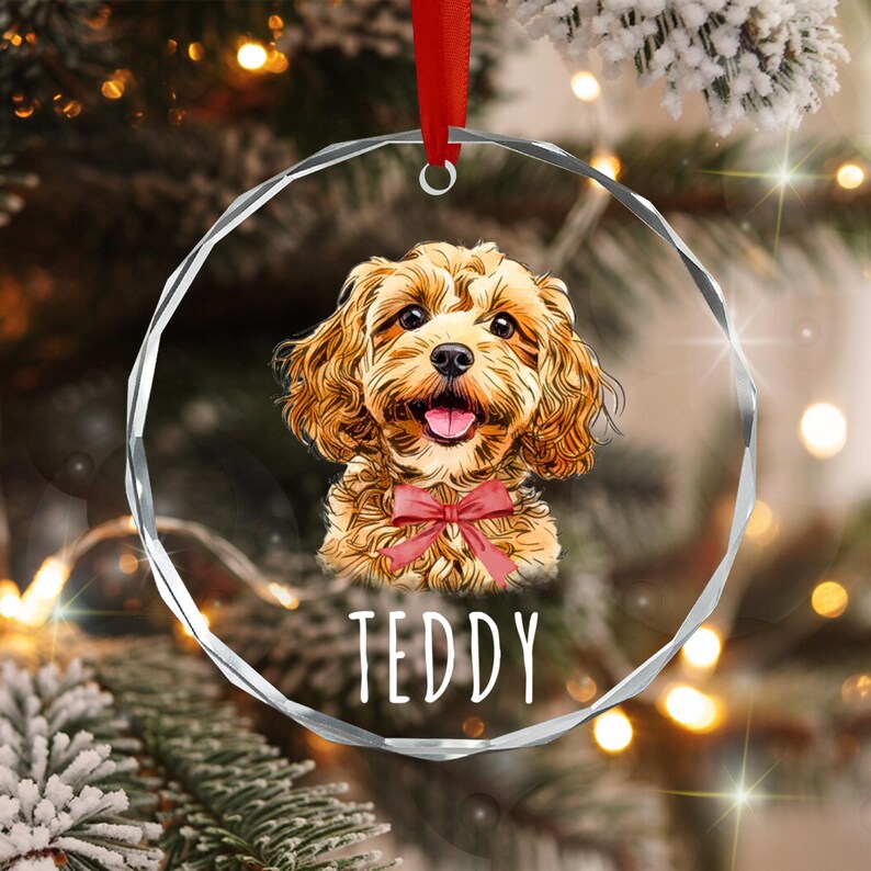 A circular, faceted glass ornament featuring a colorful illustration of a golden-brown curly-haired dog wearing a red bow tie. The name TEDDY is printed in white capital letters below the dog. A red ribbon is attached to the top.