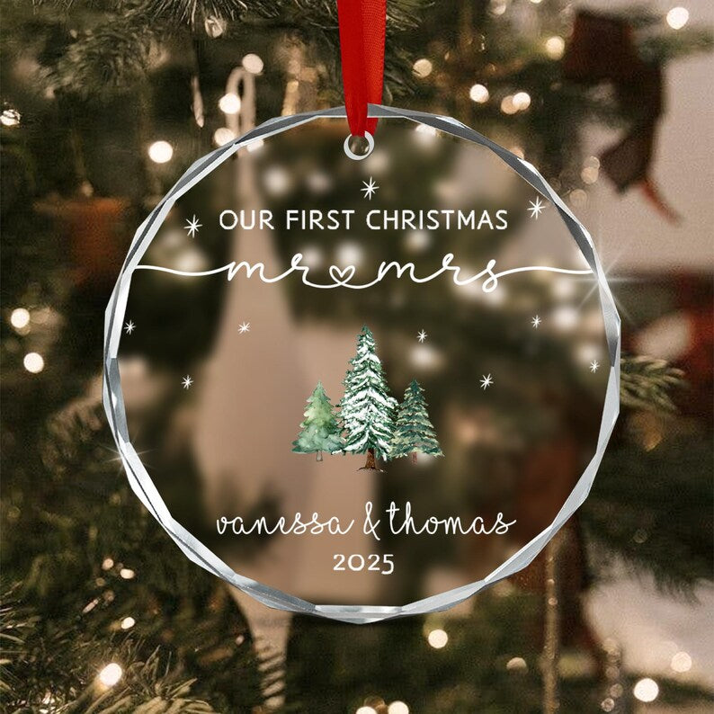 A circular clear acrylic ornament with faceted edges, featuring a red ribbon hanger. The text reads OUR FIRST CHRISTMAS mr & mrs, vanessa & thomas, and 2025. A watercolor illustration of three snow-covered pine trees is centered.