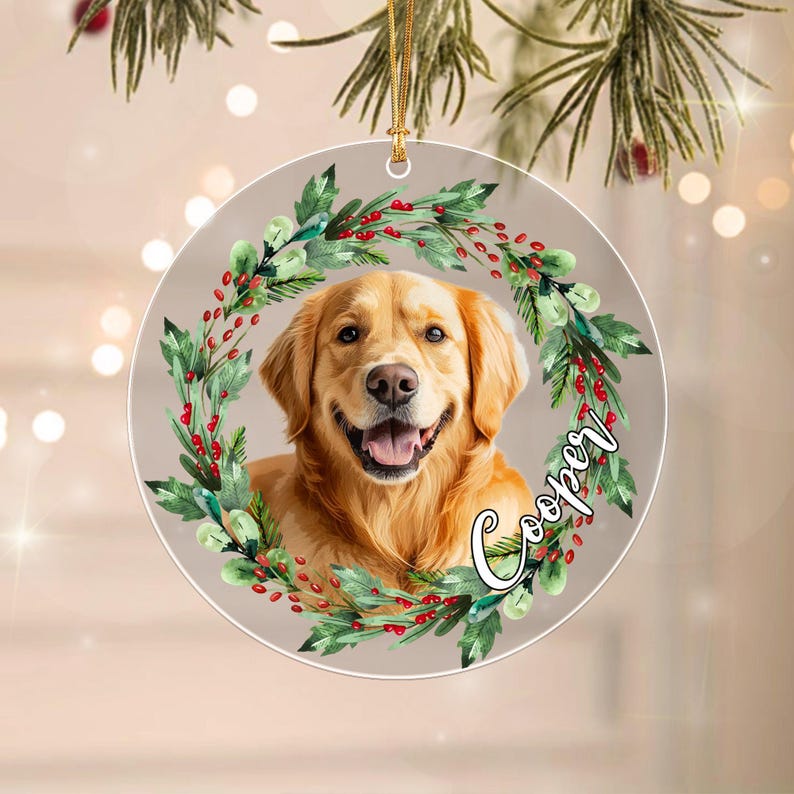 A round, flat, transparent ornament featuring a portrait of a golden retriever dog centered within a wreath of green leaves and red berries. The name Cooper is written in a white cursive script on the right side of the wreath.