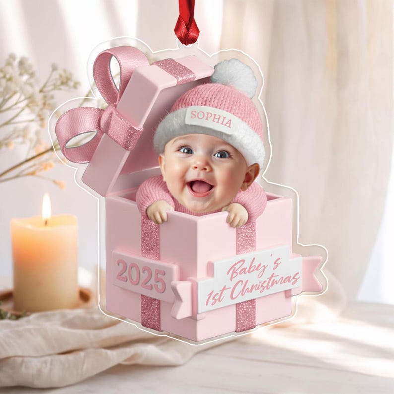 A clear acrylic Christmas ornament shaped like a pink gift box with a baby wearing a pink knit hat and sweater inside. The box features the text 2025 and Baby's 1st Christmas, with the name Sophia printed on the hat brim.
