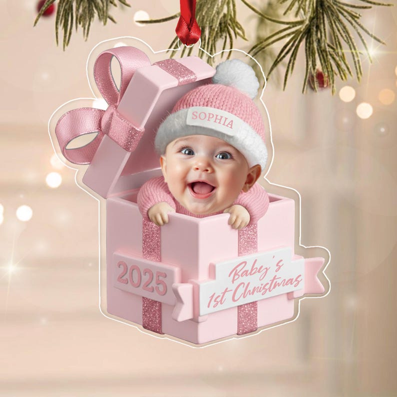 A flat, acrylic Christmas ornament shaped like a pink gift box with a large bow. Inside the box is a baby wearing a pink knit hat with a white pom-pom and the name SOPHIA. The box features the text 2025 and Baby's 1st Christmas.