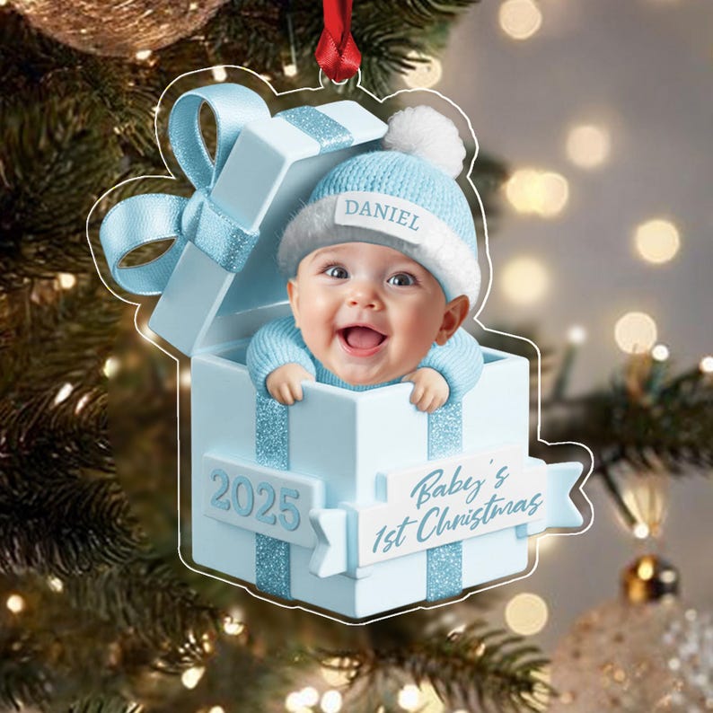 A light blue Christmas ornament shaped like a gift box with a baby wearing a matching blue knit hat with the name Daniel. The box features a ribbon, the year 2025, and the text Baby's 1st Christmas on a white banner.