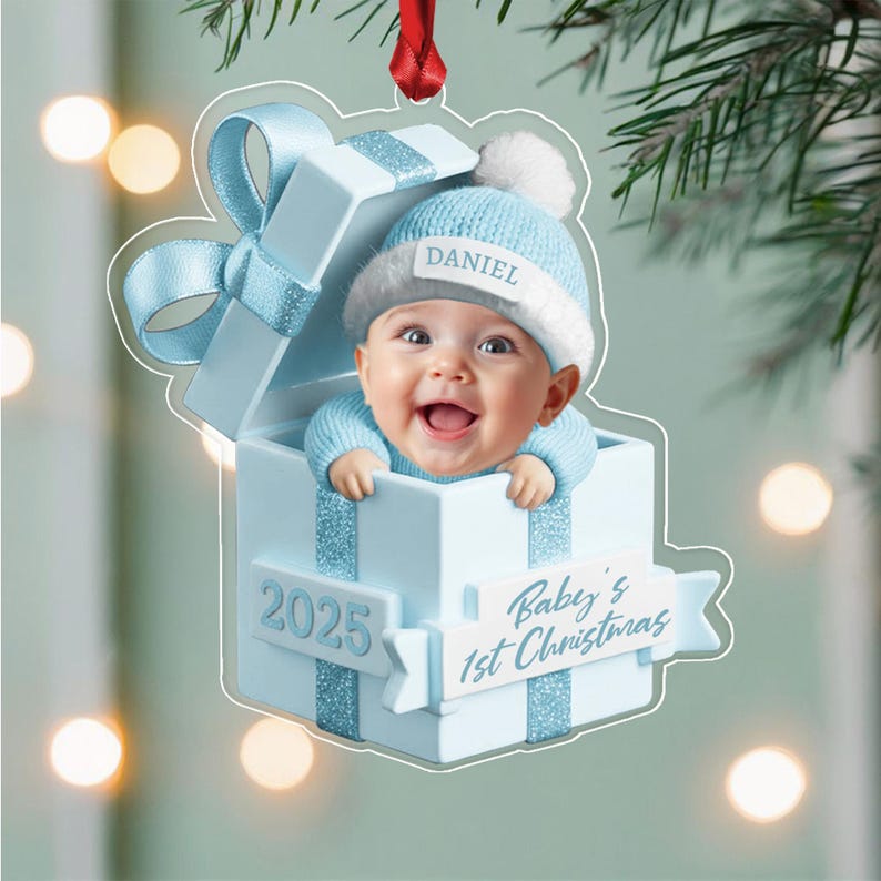 A light blue Christmas ornament shaped like a gift box with a baby wearing a matching blue knit hat with the name DANIEL printed on it. The box features the text 2025 and Baby's 1st Christmas on a white banner.