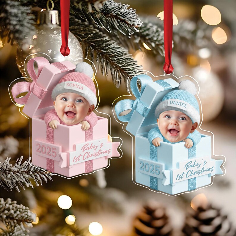 Two acrylic Christmas ornaments shaped like gift boxes, one pink and one light blue. Each features a baby wearing a matching knit hat with a name tag, 'Sophia' or 'Daniel', and a banner reading 'Baby's 1st Christmas' and '2025'.